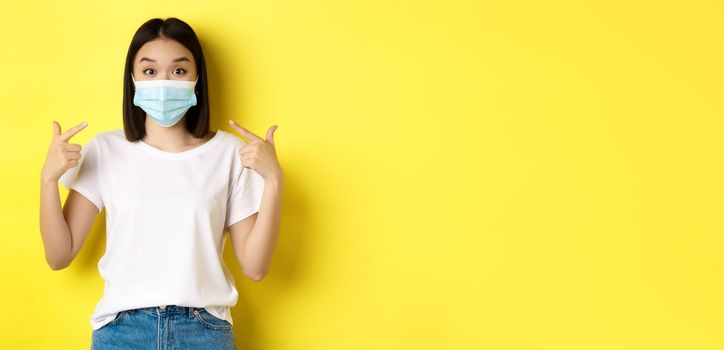 Covid-19, quarantine and social distancing concept. Young asian woman in white t-shirt, pointing at her medical mask from coronavirus, yellow background.