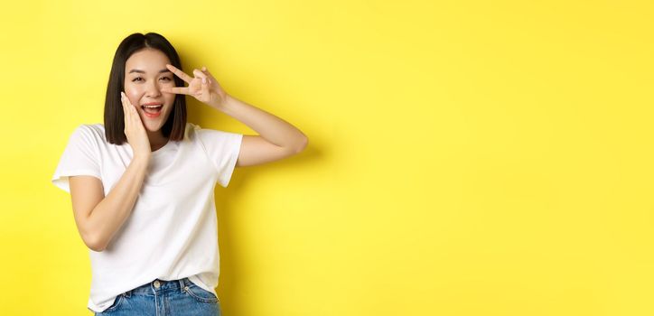 Lovely asian girl in white t-shirt posing with hand on cheek, showing peace sign on eye, standing over yellow background.