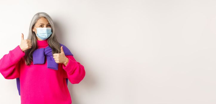 Covid, pandemic and social distancing concept. Cheerful and stylish asian senior woman wearing medical mask and showing thumbs-up, standing over white background.