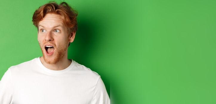 Close up of surprised and impressed redhead man checking out promotion offer, looking left with dropped jaw, standing over green background.