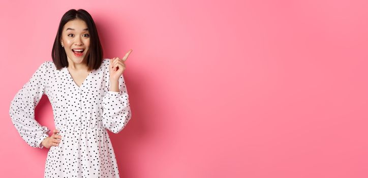 Excited pretty asian girl pointing at upper right corner, gasping amazed and looking at camera, discuss store promo offer, standing over pink background.