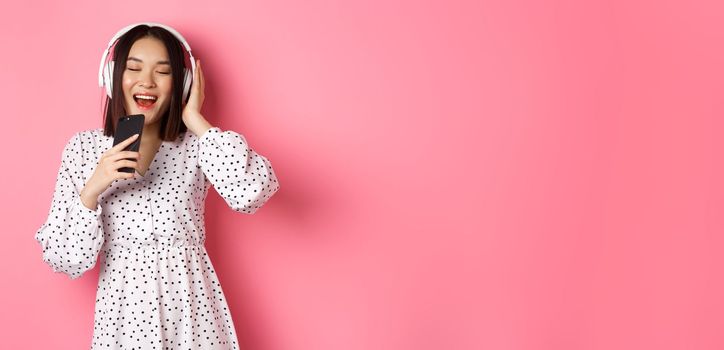 Cute asian woman playing karaoke app, singing in mobile phone and using headphones, standing in dress over pink background.