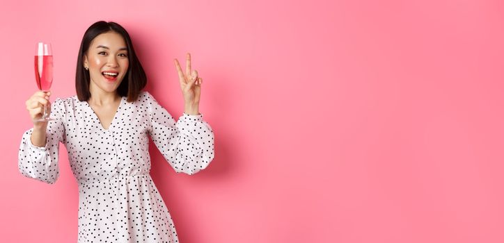 Cute asian female model drinking champagne, celebrating on party and showing peace sign, smiling happy at camera, standing over pink background.