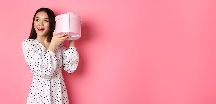 Cute asian woman shaking box with gift, smiling and looking intrigued, guess what inside present, standing over pink background.