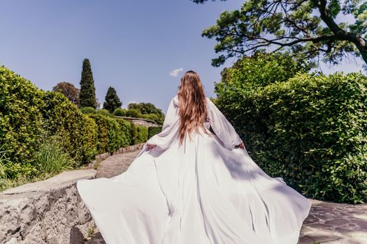 Brunette runs white dress park. A beautiful woman with long brown hair and a long white dress runs along the path along the beautiful bushes in the park, rear view.