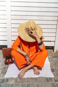 Stylish woman in an orange suit with a hat sits on a rug on a white striped background. On the hands are jewelry rings and bracelets, sandals and a bag stand side by side