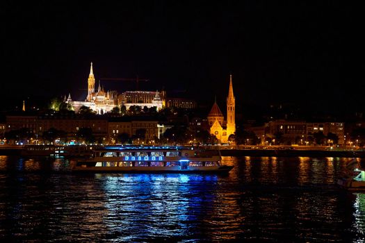 Budapest night landscape with lots of lanterns and illuminated buildings. A pleasure boat is sailing on the river. Budapest, Hungary - 08.24.2022