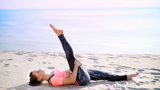 Healthy, young beautiful woman performs exercises for muscles of the press, picks up the body, legs, pumps the press lying on the sand, on the beach, at sunrise. High quality photo