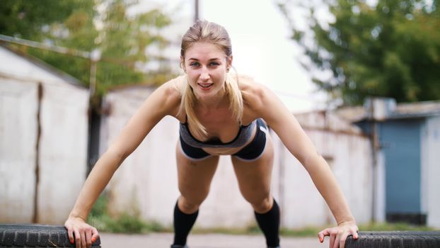 sexy athletic young blond woman in shorts, performs various strength exercises with the help of tires, push-ups, In summer, near old abandoned garages. High quality photo