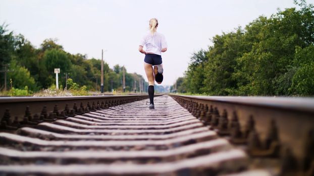 Beautiful sexy athletic young blond woman in top and shorts, running, jogging, Rear view. On the railway, on rails, sleepers, in the summer. High quality photo