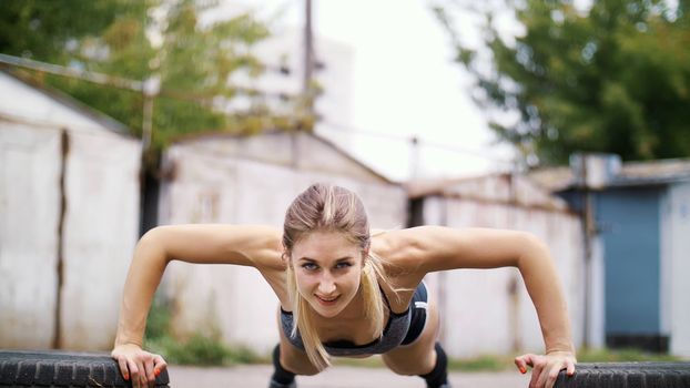 sexy athletic young blond woman in shorts, performs various strength exercises with the help of tires, push-ups, In summer, near old abandoned garages. High quality photo