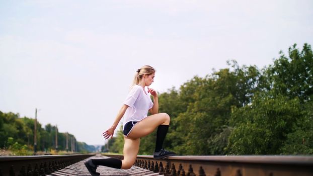 Beautiful sexy athletic young blond woman in top and shorts, performs exercises for legs, lifting legs with a bent knee, On railway, on rails, sleepers, in summer. slow motion. High quality photo