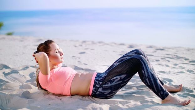Healthy, young beautiful woman performs exercises for muscles of the press, picks up the body, pumps the press lying on the sand, on the beach, at sunrise. High quality photo