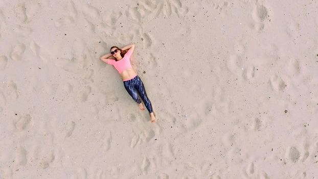 Stylish young athletic beautiful woman in sunglasses, lying on the sand, on a deserted beach, in summer. View from above, aero video. High quality photo