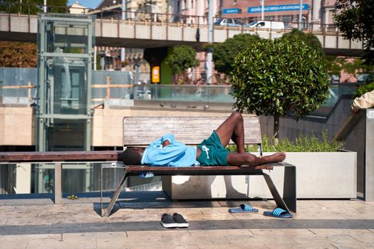 A man takes off his shoes and sleeps on a bench on the embankment in the middle of a hot summer day. Budapest, Hungary - 08.25.2022