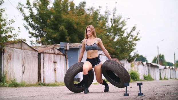 sexy athletic young blond woman in shorts, performs various strength exercises with the help of tires, lifting weights. In summer, near old abandoned garages. High quality photo