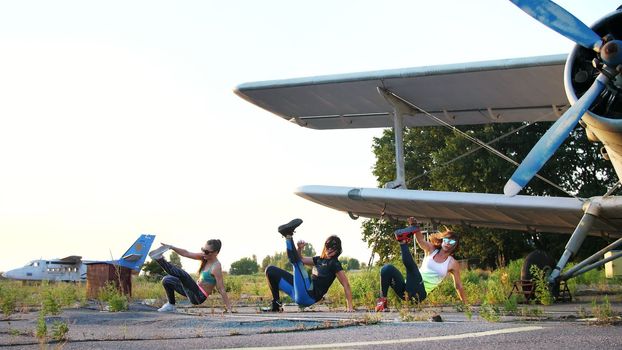 Beautiful, athletic, young women in sunglasses, in tights, perform synchronously different strength exercises, jumps, push-ups, lifting legs.on an abandoned airfield, near plane. High quality photo