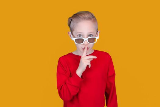 a cheerful child in children's 3D glasses puts his index finger to his lips and makes a quiet gesture