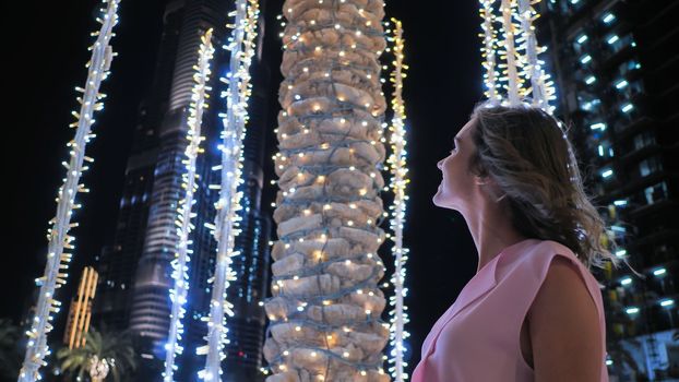The girl gently touches the glowing lights on a palm tree in the night city of Dubai