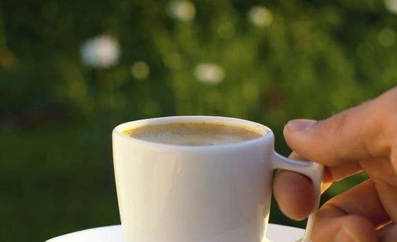 Close up male hand putting cup of coffee on a saucer on blurry nature background. Breakfast in the garden. Natural lifestyle