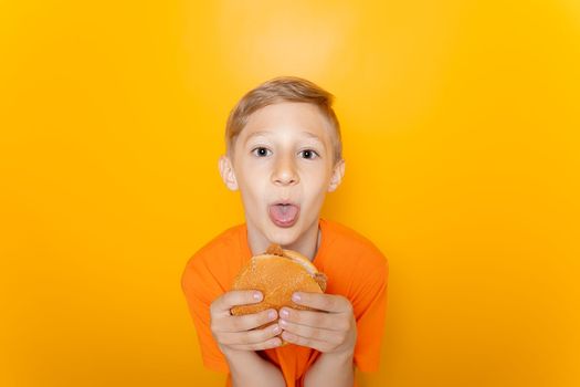 a boy in an orange T-shirt holds a hamburger in front of him and sticks his tongue out at the camera