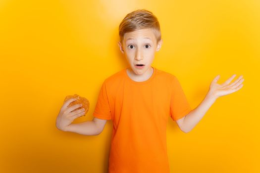 a boy in an orange T shirt holds a hamburger in one hand spreads his arms out to the sides and shows a gesture of bewilderment