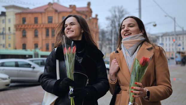 Two joyful friends in a jacket and coat are walking around the city with tulips