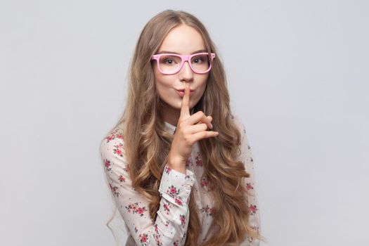 Portrait of woman in glasses with wavy blond hair showing shh gesture holding finger near lips, with naughty smile on face, making surprise, secret. Indoor studio shot isolated on gray background.