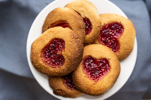 Delicious warm Homemade cookies on white plate, decorated with raspberry jam in the shape of a heart. View from above. selective focus. Top view of traditional Christmas cookies filled with red jam. Traditional filled biscuits