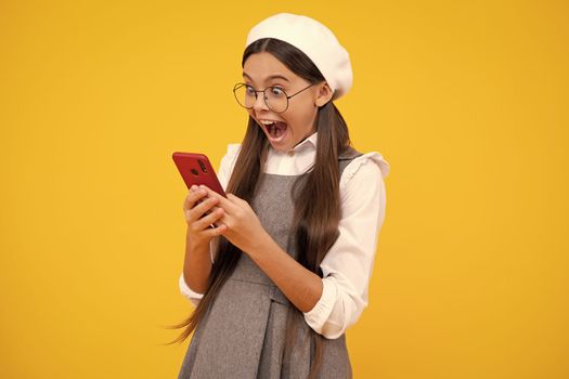 Excited teenager girl use smart phone, share social media, chatting by mobile phone wear stylish casual trendy clothes isolated over yellow background. Child with smartphone