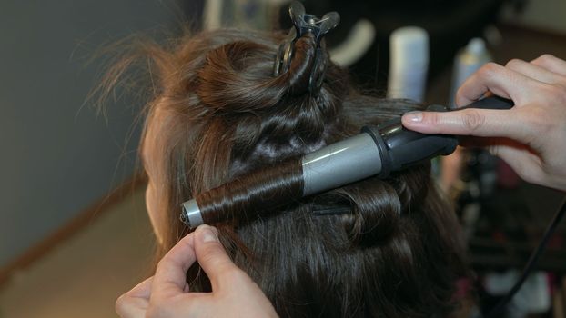The hairdresser curls the hair of a girl in a beauty salon using a curling iron