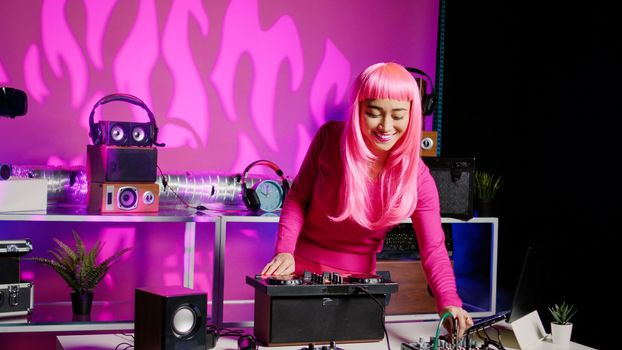 Smiling musician working as dj playing at mixer console, mixing techno sound with eletronic using audio equipment. Performer with pink hair having fun performing in club at night time