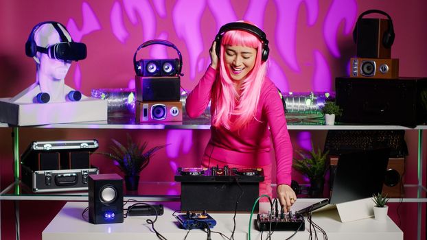 Smiling dj woman dancing and mixing techno sound with electronic remix using professional turntables, having fun during concert in nightclub. Asian artist standing over pink background in studio