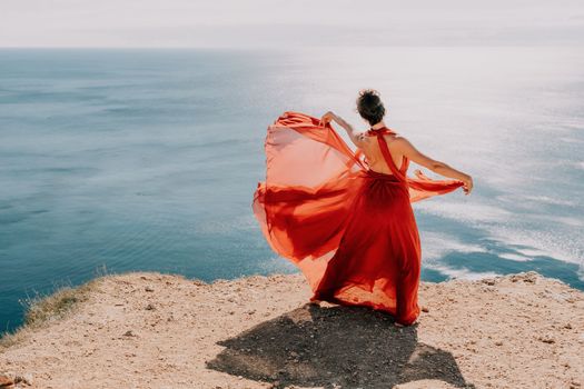 Side view a Young beautiful sensual woman in a red long dress posing on a rock high above the sea during sunrise. Girl on the nature on blue sky background. Fashion photo.