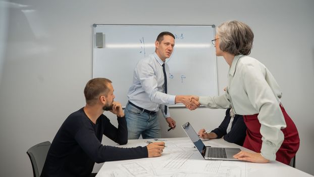 The boss makes a presentation to subordinates at the white board. Caucasian man shaking hands with middle aged woman