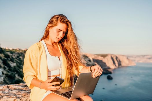 Successful business woman in yellow hat working on laptop by the sea. Pretty lady typing on computer at summer day outdoors. Freelance, travel and holidays concept.