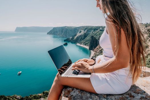 Successful business woman in yellow hat working on laptop by the sea. Pretty lady typing on computer at summer day outdoors. Freelance, travel and holidays concept.