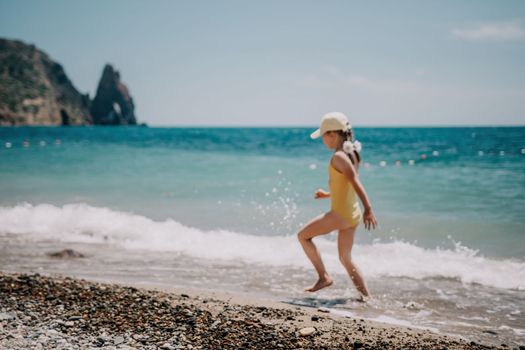 Cute little girl running along the seashore against a clear blue sea and rejoices in the rays of the summer sun. Beautiful girl in yellow swimsuit running and having fun on tropical beach