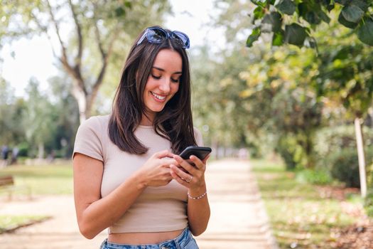 smiling young caucasian woman typing on her mobile phone in a park, concept of youth and technology of communication