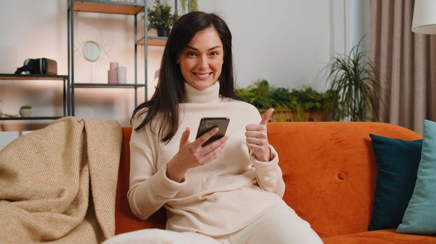 Happy brunette young woman use mobile smartphone typing browsing say Wow yes found out great big win good news celebrate lottery jackpot doing winner gesture. Girl at home in room sitting on sofa