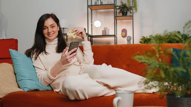 Young brunette woman using credit bank card and smartphone while transferring money, purchases online shopping, order food delivery at home apartment indoors. Happy girl in living room lying on couch