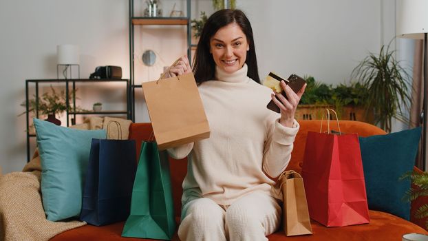 Happy shopaholic consumer young woman sitting with shopping bags at home apartment sitting on couch making online order, payment with credit plastic bank card. Discount sale on internet store mall