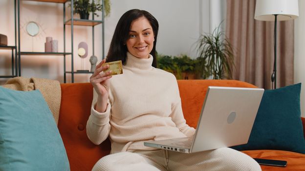 Young woman using credit bank card and laptop computer while transferring money, purchases online shopping, order food delivery at home apartment. Adult girl in day living room sitting on couch indoor