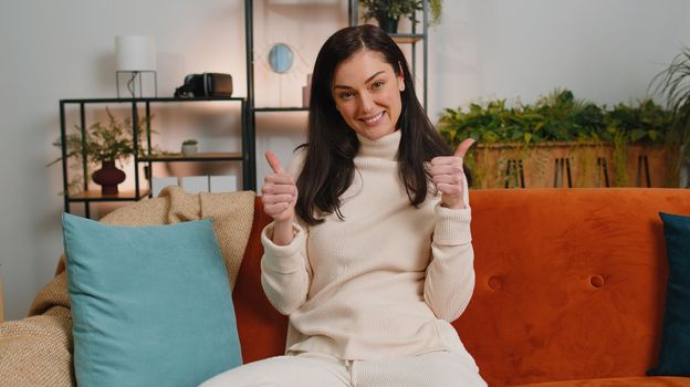 Like. Happy excited brunette woman looking approvingly at camera showing thumbs up, like sign positive something, good great news, positive feedback. Adult girl sitting on couch at home living room