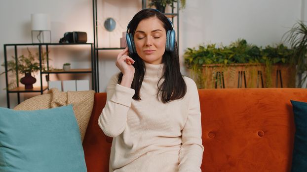 Happy overjoyed brunette woman in wireless headphones relaxing sitting on couch at home apartment choosing listening favorite energetic disco dancing music. People weekend daytime leisure activities