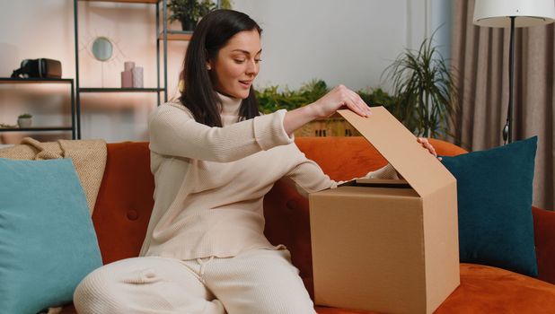 Happy surprised winner young woman unpacking delivery parcel at home. Smiling satisfied girl shopper online shop customer opening cardboard box receiving purchase gift present by fast postal shipping