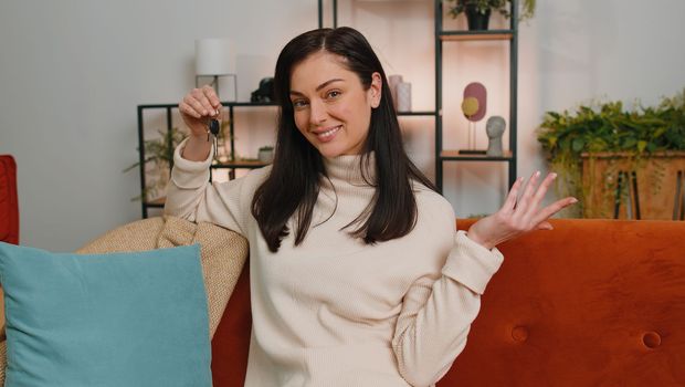 Young woman real estate agent lifting hand showing the keys of new home house apartment, buying or renting property, mortgage loan at modern home apartment indoors. Adult girl in room sitting on sofa