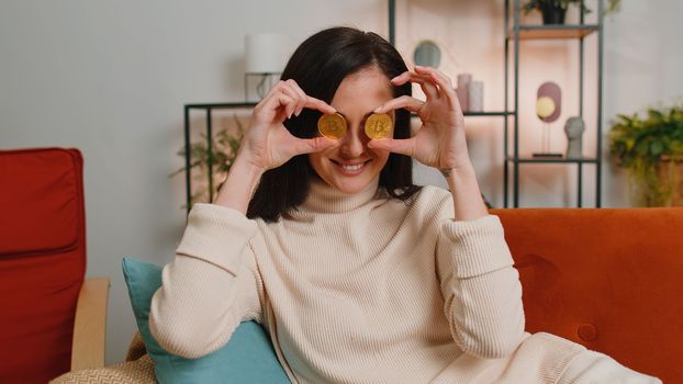 Woman freelancer holding two gold coins at home, successful developer programmer. Girl stock trader earning bitcoins after online monitoring trading operations. Increasing wealth, financial prosperity