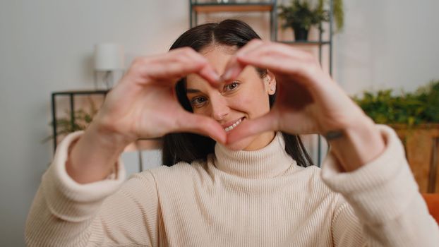 I love you. Happy brunette woman at home living room couch makes symbol of love, showing heart sign to camera, express romantic feelings express sincere positive feelings. Charity, gratitude, donation