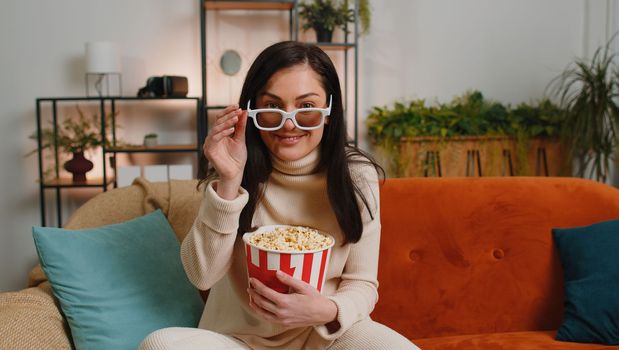 Excited woman in 3D glasses sits on sofa eating popcorn snacks and watching interesting TV serial, sport game, film, online social media movie content at home apartment. Girl enjoying entertainment